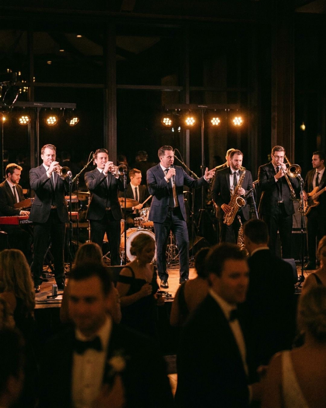 A nine-piece event band with a full horn section performing on a warmly lit stage while guests fill the dance floor at a wedding reception
