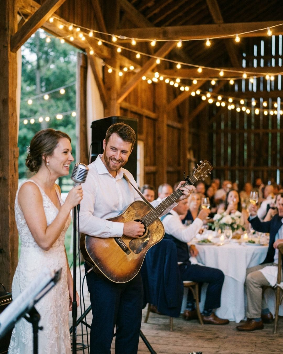 An acoustic duo, a female singer and a male guitarist, performing at a barn wedding reception under string lights while seated guests watch