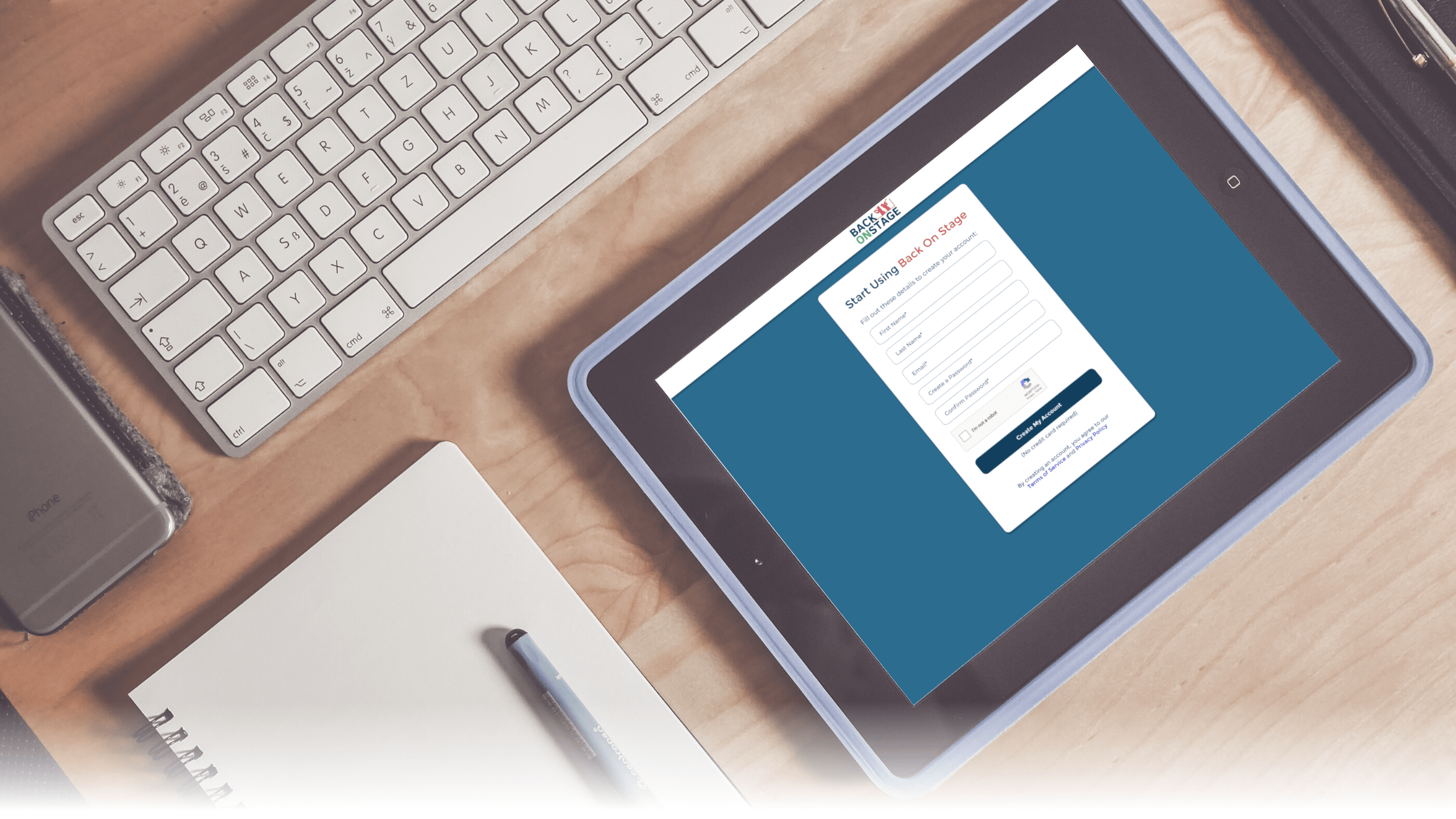 An iPad on a wooden desk showing the Back On Stage account creation screen, beside a keyboard, phone and notebook
